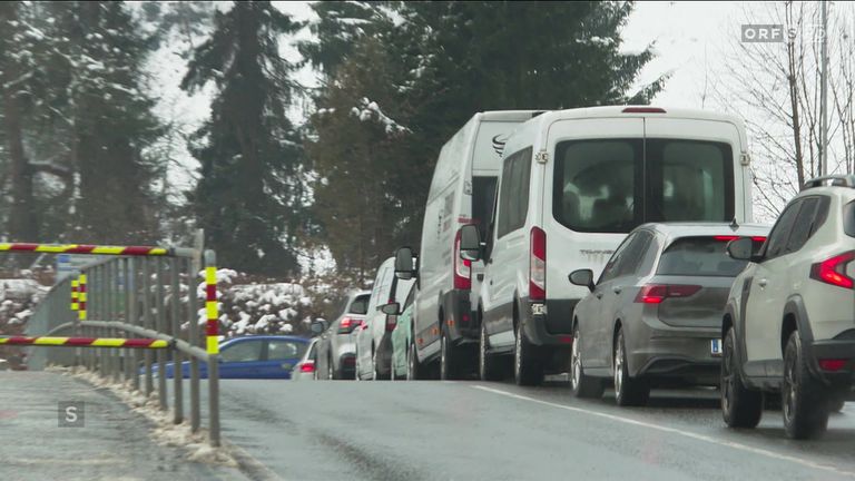 A line of cars and vans are parked on a road, with trees covered in snow in the background.