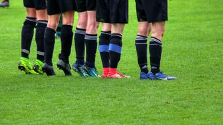 Soccer players in different colored jerseys stand on a grass field, possibly preparing for a match. The players' feet are clad in sneakers, some with visible brand logos.