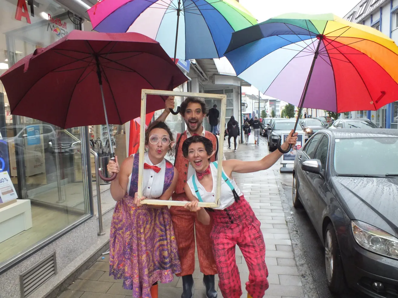 Three people dressed in costumes pose with umbrellas on a sidewalk. One person holds a picture frame, and cars are parked on the road.
