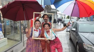 Three people dressed in costumes pose with umbrellas on a sidewalk. One person holds a picture frame, and cars are parked on the road.