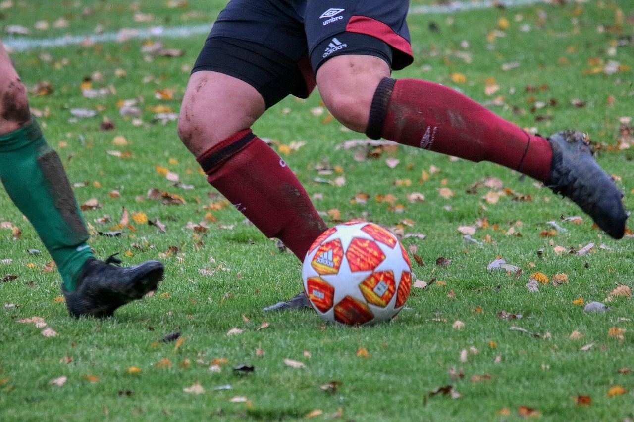 A soccer player is kicking a soccer ball on a grass field with fallen leaves, wearing red and black socks.