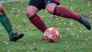 A soccer player is kicking a soccer ball on a grass field with fallen leaves, wearing red and black socks.