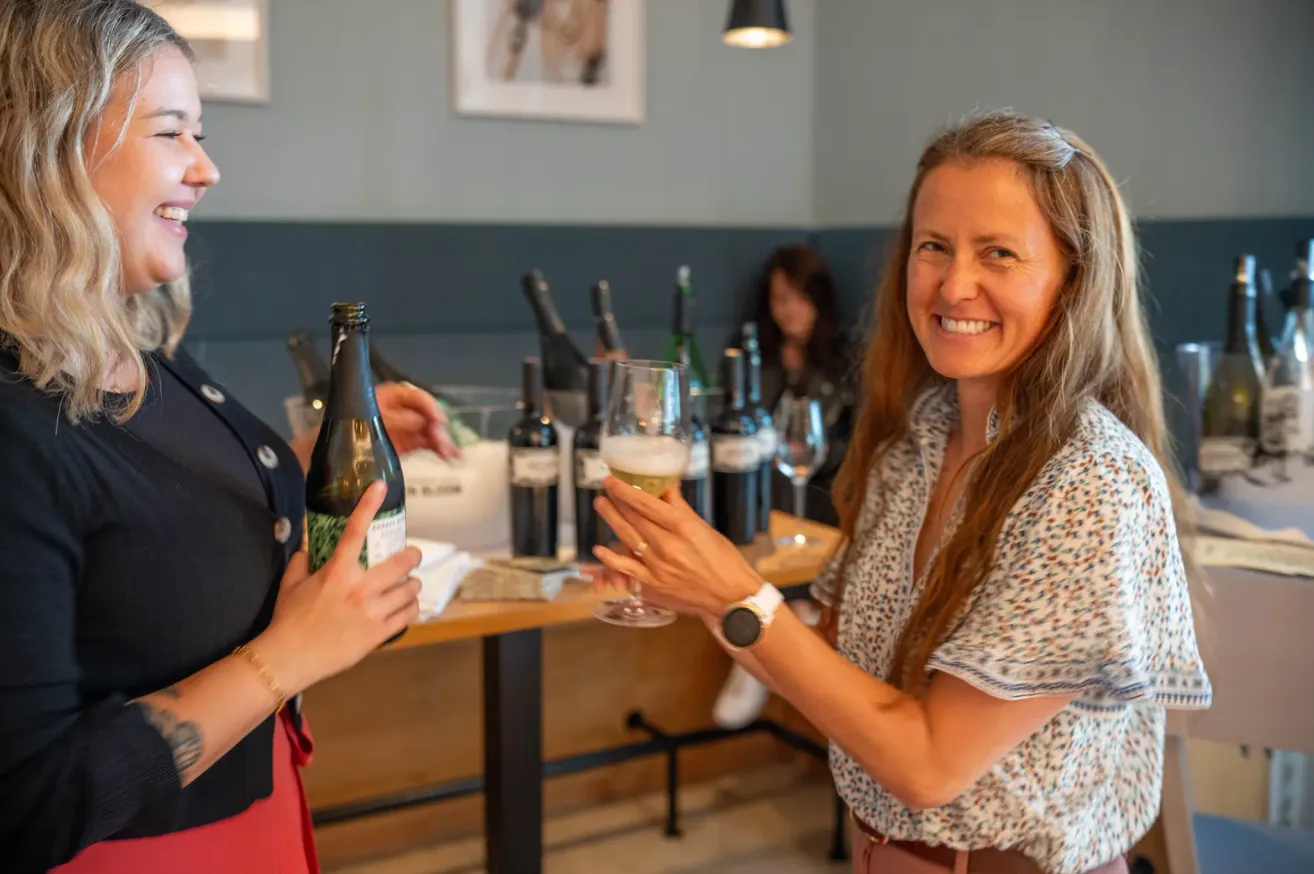 Two women in a restaurant toasting with a glass of sparkling wine. Behind them, wine bottles are displayed on a table.