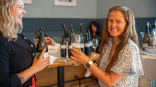 Two women in a restaurant toasting with a glass of sparkling wine. Behind them, wine bottles are displayed on a table.
