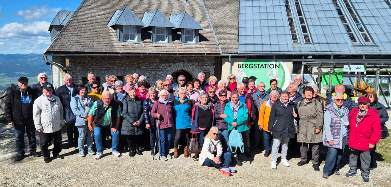 A group of elderly people, possibly tourists, pose for a photo outside a building with a sign reading Bergstation.
