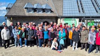 A group of elderly people, possibly tourists, pose for a photo outside a building with a sign reading Bergstation.