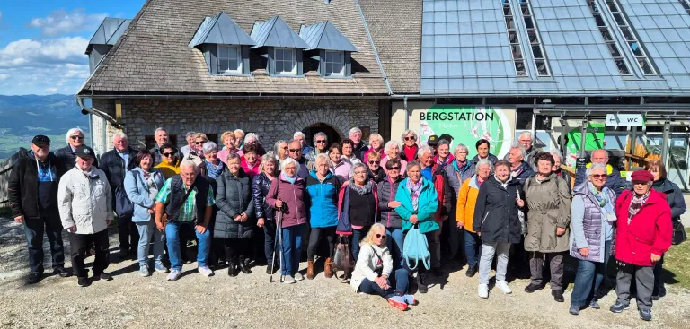 A group of elderly people, possibly tourists, pose for a photo outside a building with a sign reading Bergstation.
