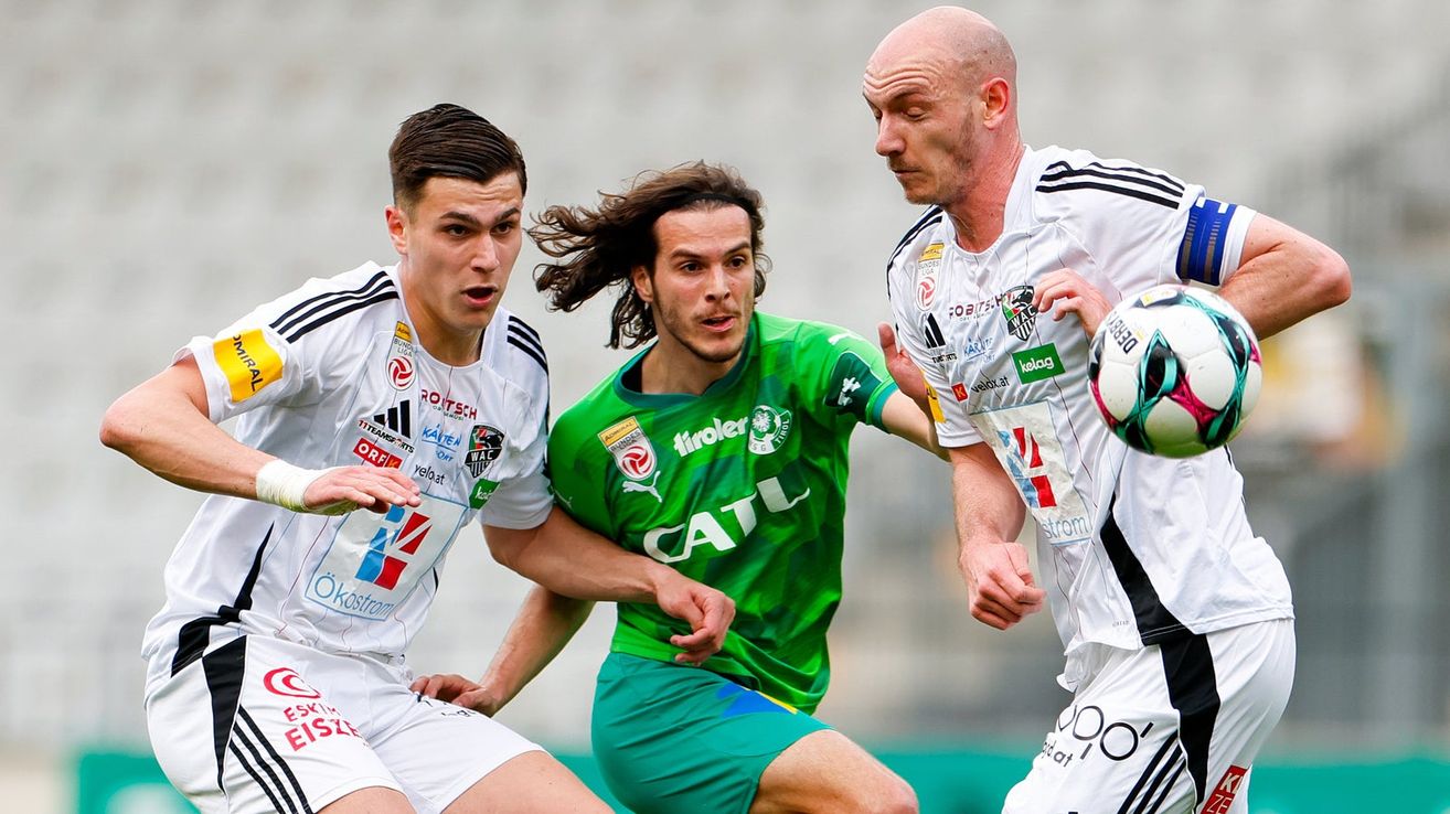 Three soccer players in action. One in green, another in white, and one bald player also in white. The green player is being pushed by the bald player.