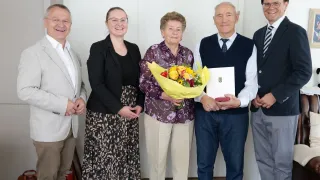 Five people stand together, smiling. The woman in the center holds a bouquet of flowers and a certificate. The man next to her wears a vest. The others wear suits and glasses.
