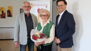 Three individuals stand in front of a banner. The woman holds a bouquet of flowers, and the two men wear formal attire. They appear to be at an event.