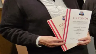 A man holds a certificate in his hands. He wears a black jacket and jeans. The certificate has a red border. He stands in front of a brown box with the word 'Filmen' written on it.