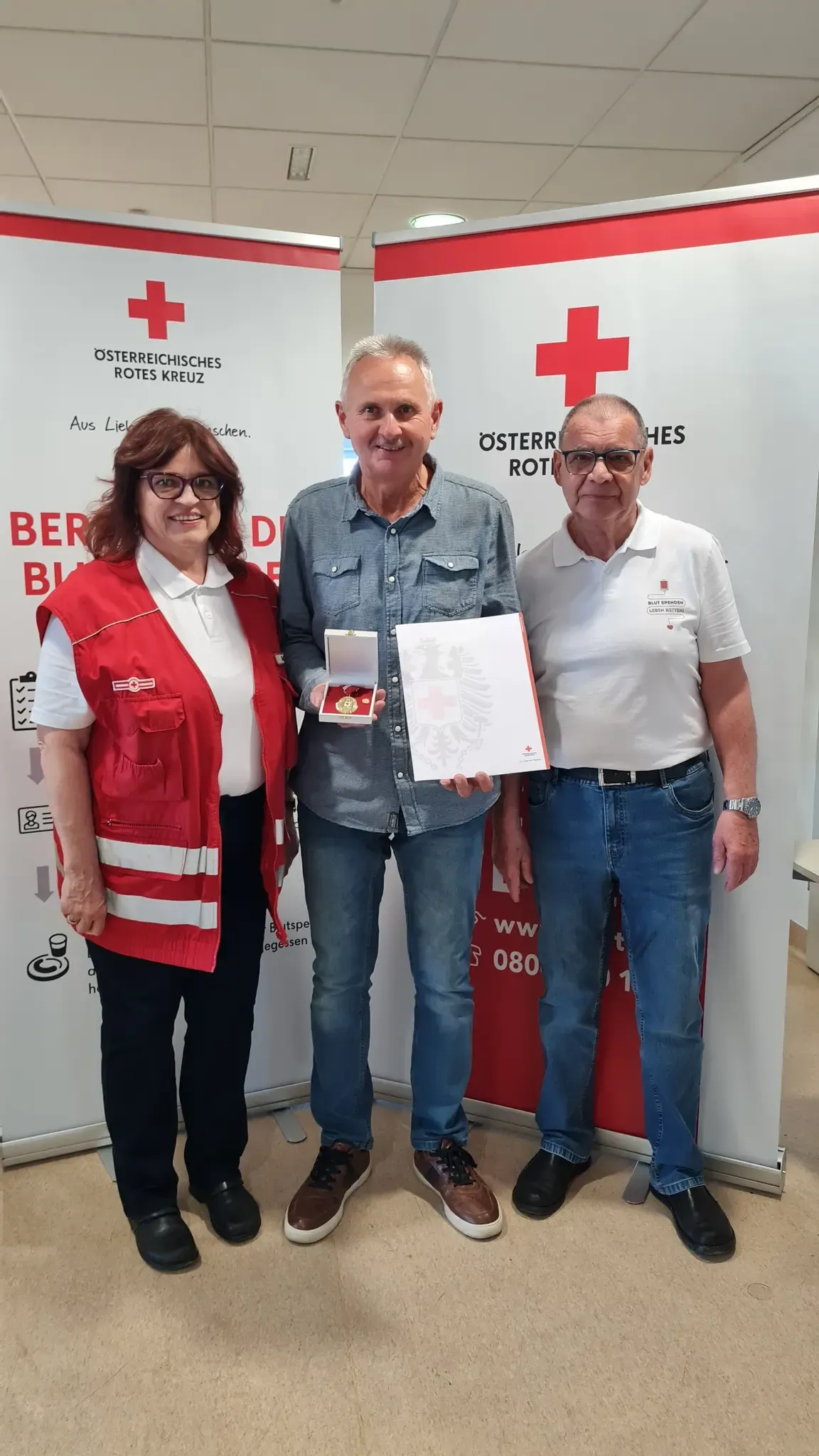 Three individuals stand near a wall with the Red Cross logo. A woman and two men. They are smiling and appear to be posing for a photo.