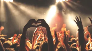 A large crowd at a concert forms a heart shape with their hands. The stage is illuminated with spotlights.