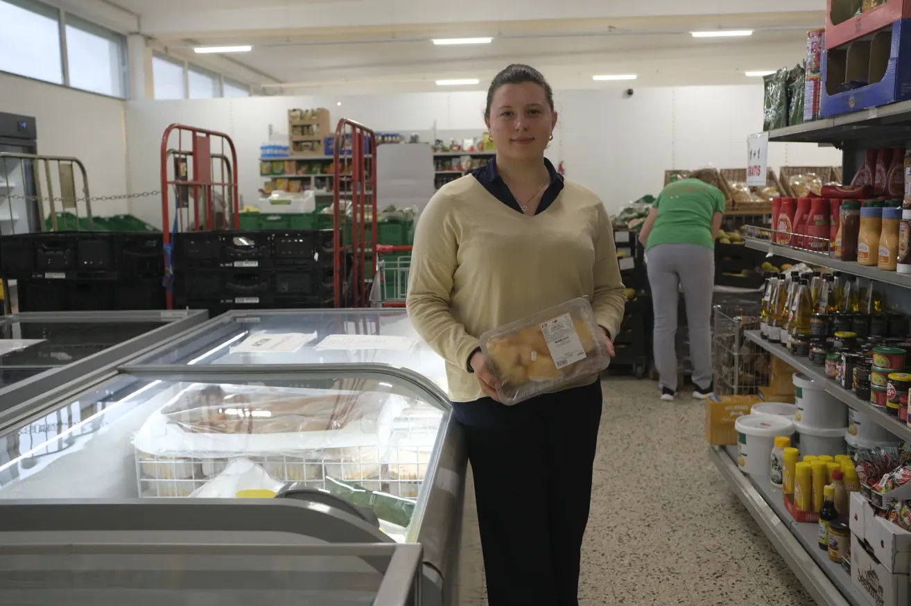 A woman in a yellow sweater holds a package of food in a supermarket. Behind her, a freezer is visible with fish inside. Another person in a green shirt is in the background.
