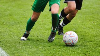 Two soccer players on a grassy field. One in green and one in black. The green player is about to kick the ball.