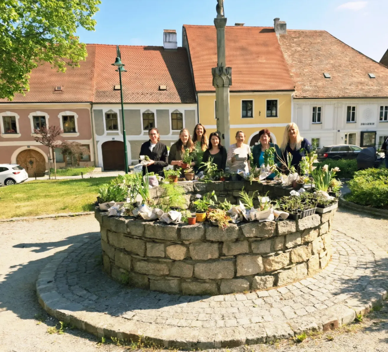 A group of women stand around a fountain with potted plants. They are in front of a building with red roofs and a statue.