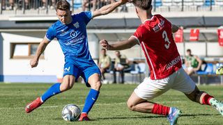 Two soccer players, one in blue and one in red, are playing on a field. One is preparing to kick the ball while the other tries to block.