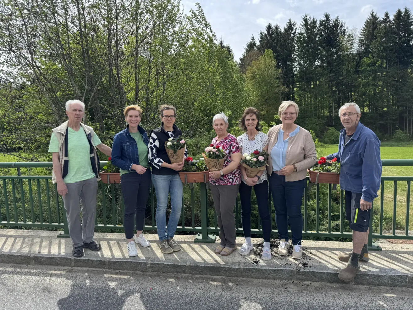 A group of elderly individuals pose for a photo while holding flower pots, standing on a concrete surface with a green railing in front of them. They are smiling and seem to be posing for a photo.