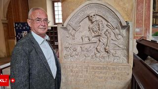 An older man stands next to a monument with a skeleton figure and an inscription. The man wears glasses and a gray jacket. The background shows a wooden shelf.