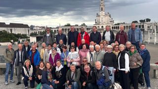A group of people in casual clothing stand in front of a building with a tower. Some wear glasses and scarves. The sky is cloudy.