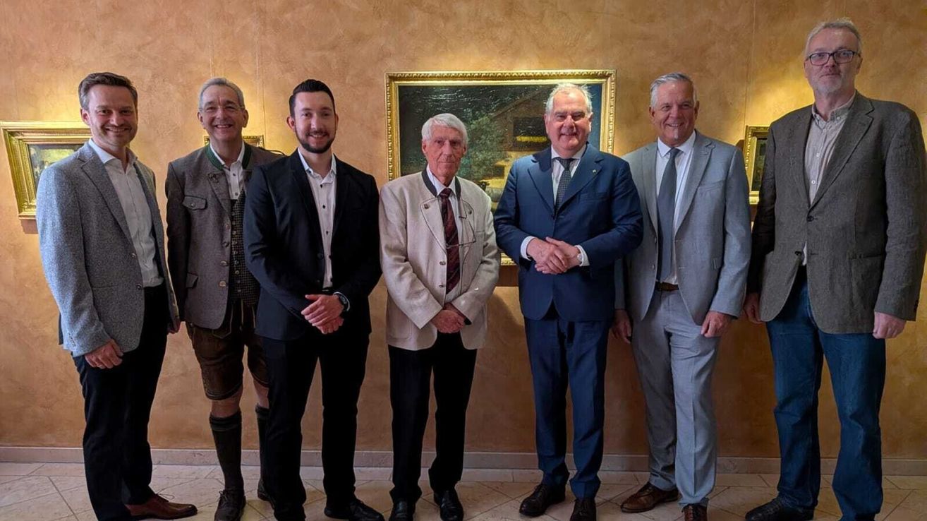 Fünf Männer in Anzügen stehen vor einer Wand mit einem Gemälde. Sie scheinen für ein Foto zu posieren.