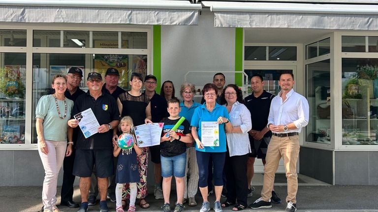 A group of people, including adults and children, stand together outside a building, holding certificates and smiling for a photo.