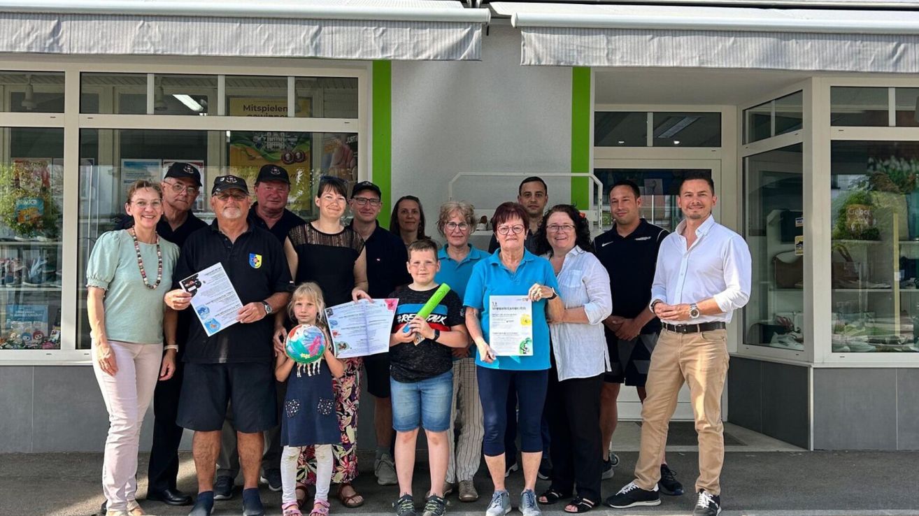 A group of people, including adults and children, stand together outside a building, holding certificates and smiling for a photo.