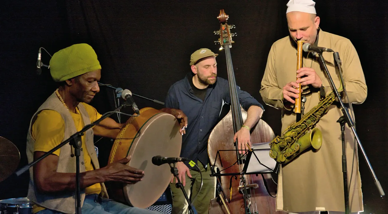 Drei Männer spielen auf der Bühne mit Musikinstrumenten. Einer spielt eine Trommel, ein anderer ein Saxophon und der dritte einen Bass. Alle tragen Freizeitkleidung und scheinen konzentriert auf ihre Aufführung.