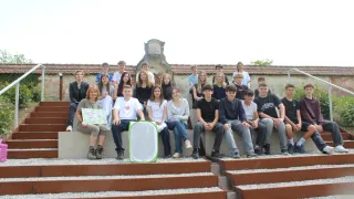 Eine Gruppe von Teenagern sitzt auf Betonstufen und posiert für ein Foto. Eine Frau hält ein Bild eines Vogels. Vor der Gruppe steht ein grünes Netz.