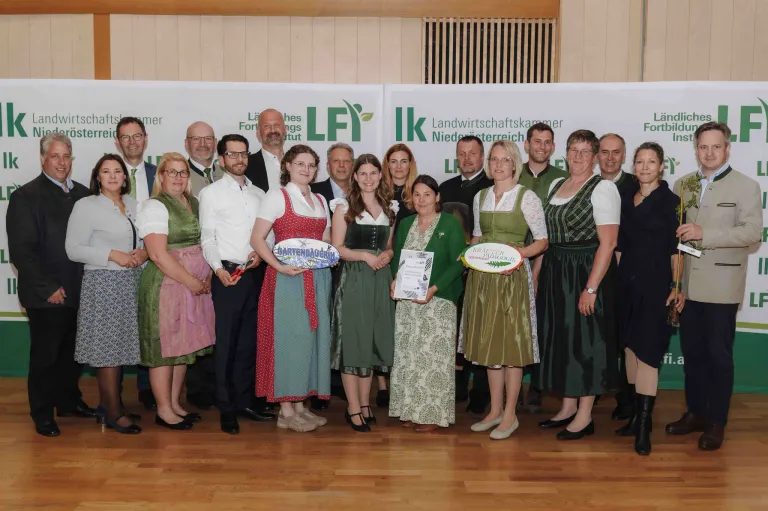 A group of people are posing for a photograph with some holding trophies and certificates. They are dressed in traditional costumes. The backdrop has banners with logos.