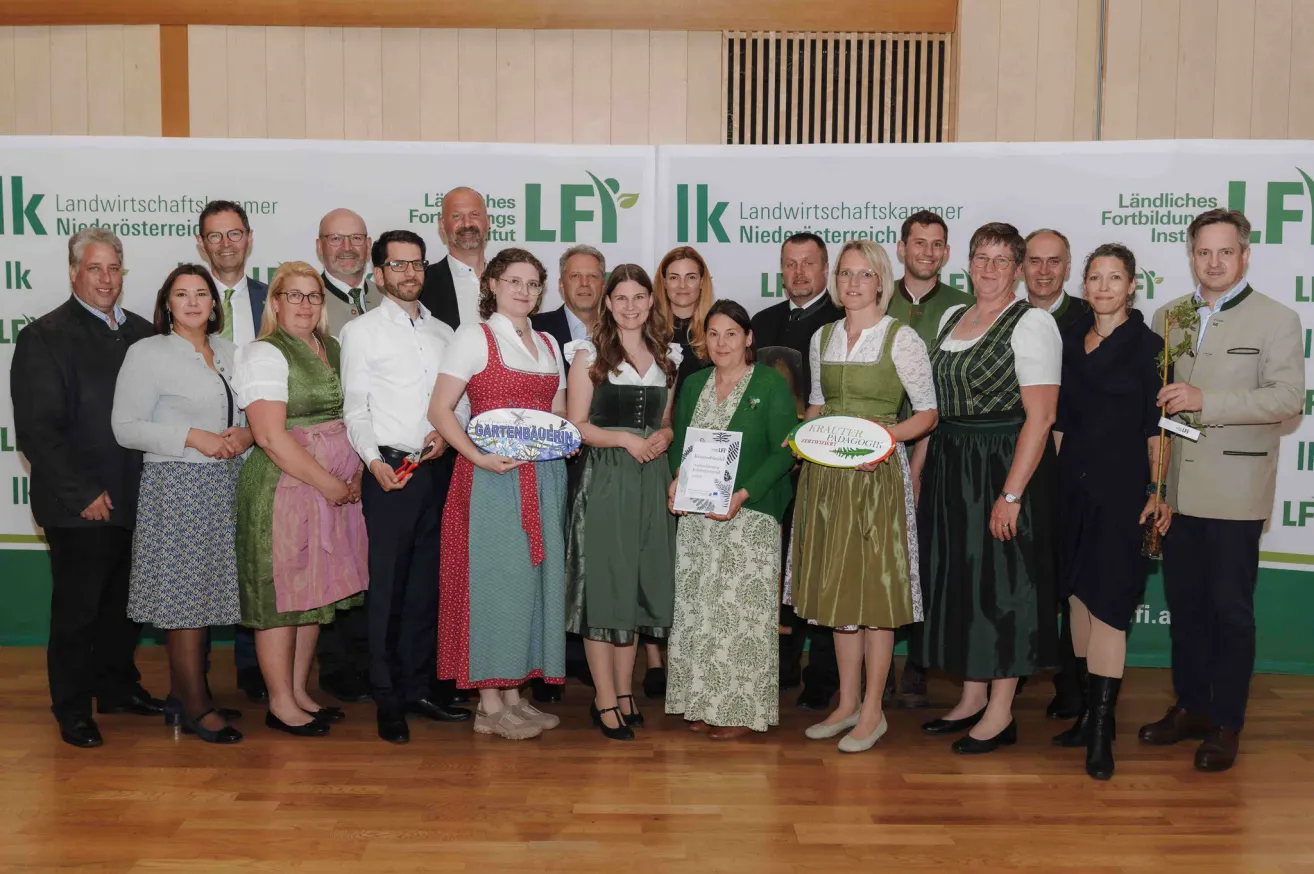 A group of people are posing for a photograph with some holding trophies and certificates. They are dressed in traditional costumes. The backdrop has banners with logos.