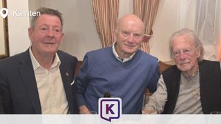 Three men are seated at a table, smiling for a picture. The man on the left wears a suit, the middle one a blue sweater, and the right one has glasses. Behind them is a curtain.