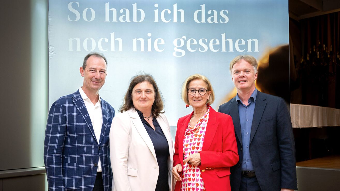 Four adults stand in front of a banner with text in German. They all smile and look at the camera. The man on the left wears a blue checkered blazer.
