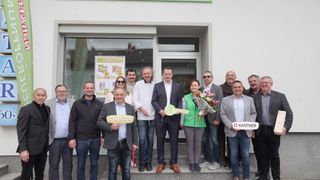 A group of people stand in front of a store. They hold signs and a key, and one woman holds a bouquet. A man holds a green sign with the text Nah&Frisch.
