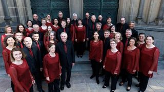 A choir in red dresses and ties stands on steps in front of a large green door. They are smiling and posing for a photo.