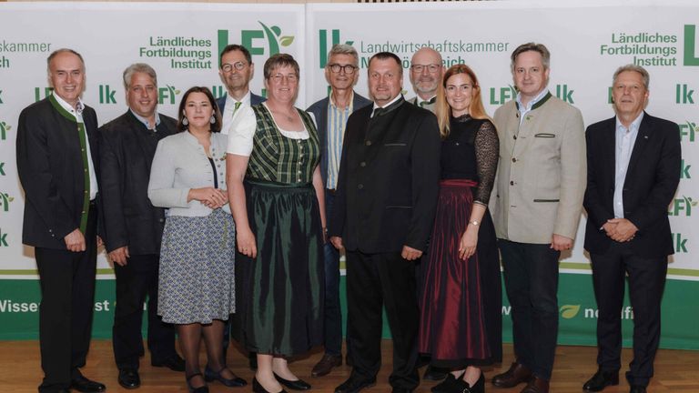 A group of people are standing and posing for a photo in front of a banner with 'LF' and 'Landwirtschaftskammer' written on it. They are wearing formal attire.