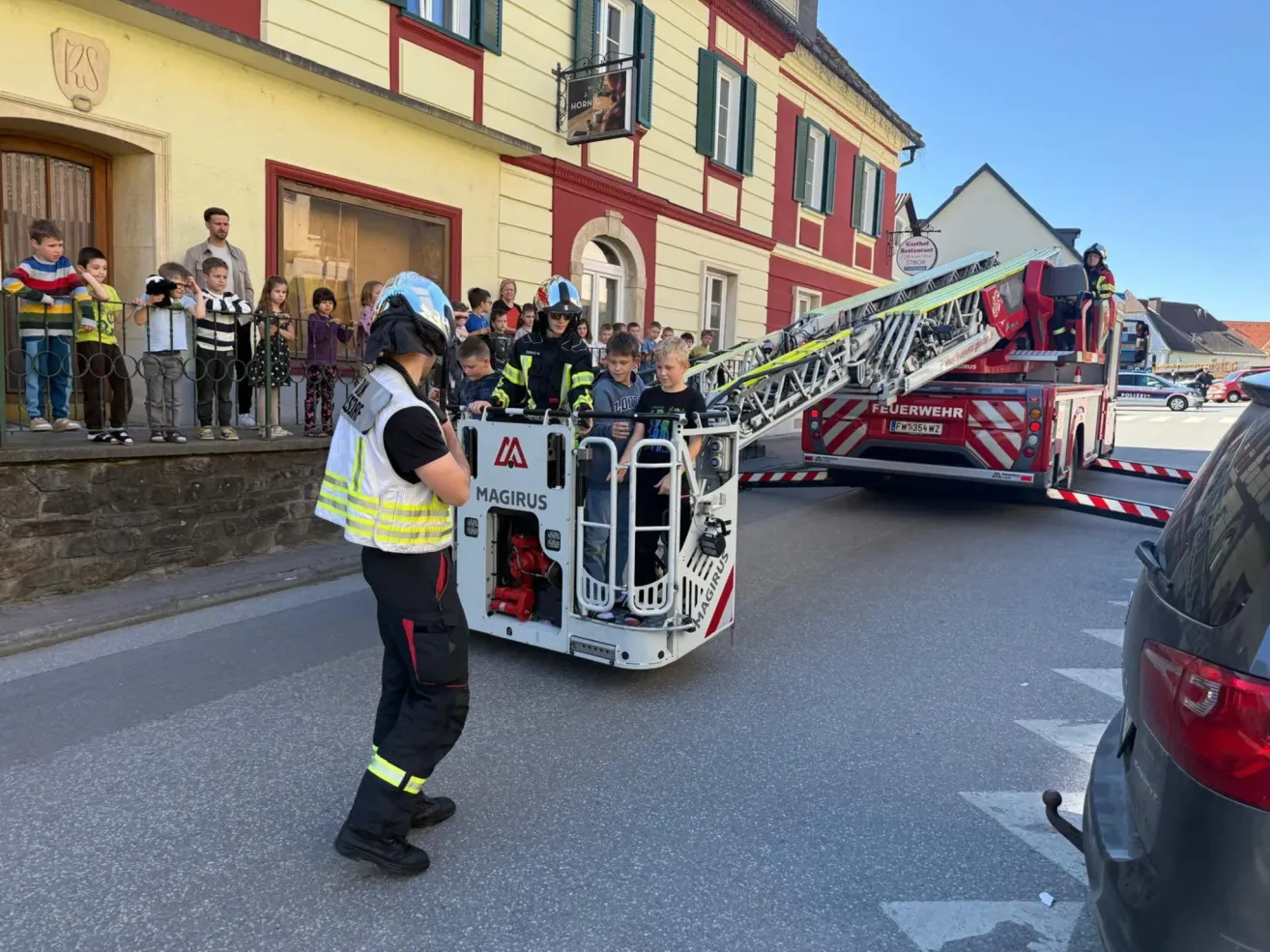Firefighters are standing in front of a building. One firefighter is letting kids experience the ladder truck.