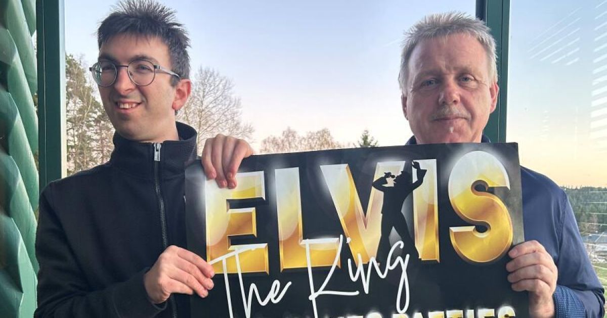Two men stand outside holding a sign that says Elvis the King with a silhouette of Elvis in a black and gold color scheme.
