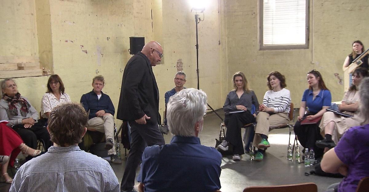 A group of people in a room, some seated and others standing, listening to a man speaking. The room has a yellow wall and a window with blinds.