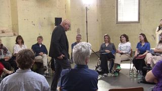 A group of people in a room, some seated and others standing, listening to a man speaking. The room has a yellow wall and a window with blinds.