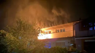 A building is on fire at night, with smoke rising into the sky. A car is parked in front, partially visible.
