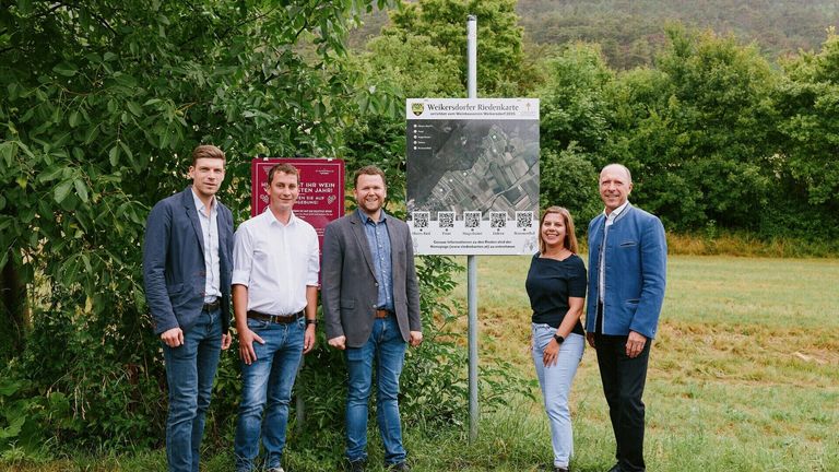 Five people stand outside near a sign with a map, smiling at the camera. Behind them, a lush, green forest and a clear sky.