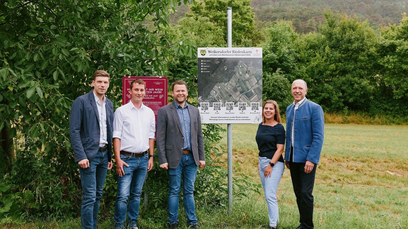 Five people stand outside near a sign with a map, smiling at the camera. Behind them, a lush, green forest and a clear sky.