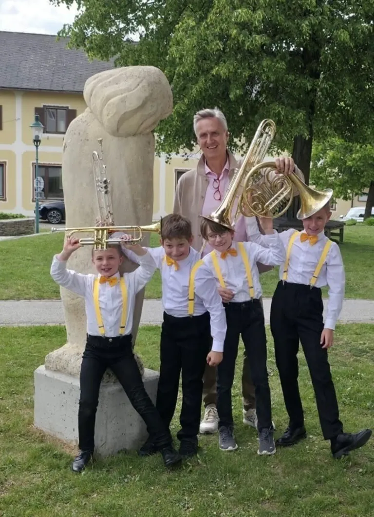 Vier Jungen in identischen Uniformen halten Musikinstrumente in der Hand und stehen vor einer großen Statue und einem lächelnden Mann, mit einem Gebäude und Bäumen im Hintergrund.