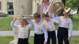 Four boys in matching uniforms hold musical instruments, standing in front of a large statue and a smiling man, with a building and trees in the background.