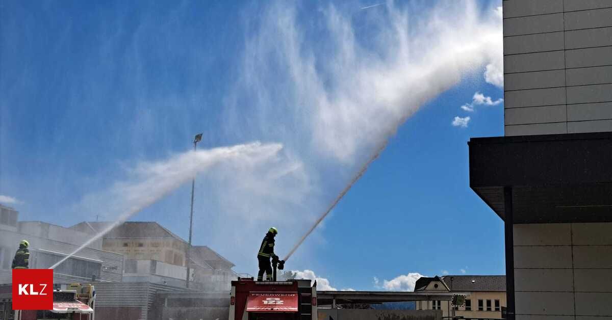 Ein Feuerwehrmann steht auf einem roten Feuerwehrwagen und sprüht Wasser aus einem Schlauch. Eine große Wasserwolke ist sichtbar. Gebäude und eine Straßenlaterne sind im Hintergrund.