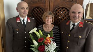 Three individuals in formal attire stand together in front of a wooden lattice backdrop. The woman holds a bouquet of flowers and wears a necklace. The men wear uniforms with red and gold emblems.