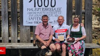 Three people and a dog sit on a bench in front of a sign that reads 1000. The man holds a cake.