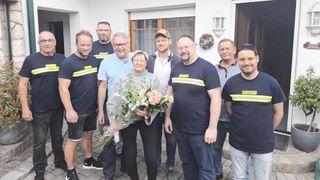 A group of men in uniforms stand behind a woman holding flowers, likely celebrating an event. They are all smiling and posing for a photo.