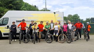 A group of cyclists, wearing helmets and cycling attire, pose for a photo in front of a van. The van has text and a logo on its side. Trees and a fence are in the background.